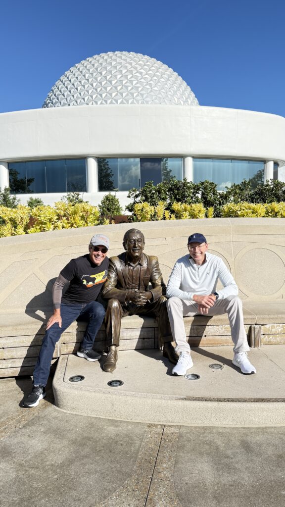 two men sitting next to Walt Disney statue
